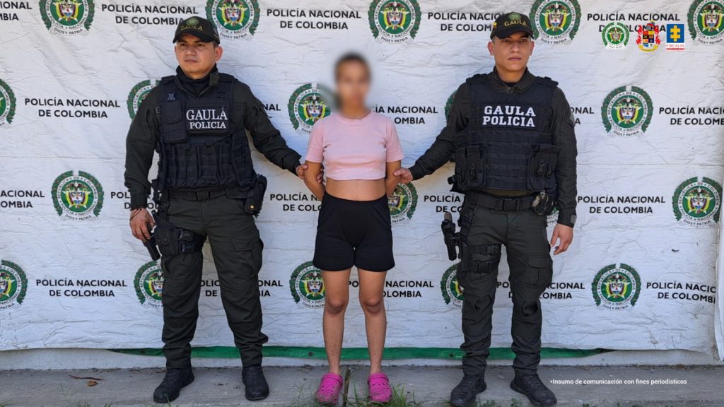 en la imagen se ve una mujer detenida bajo custodia de dos integrantes de la Policía. Detrás suyo un backing institucional.