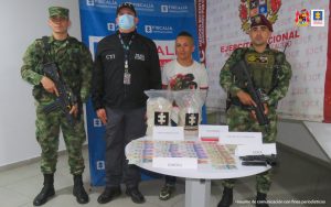 En la imagen se ve una persona detenida bajo custodia del Ejército y el CTI. Detrás suyo backings institucionales, y delante de ellos una mesa con base de coca y otros elementos incautados.