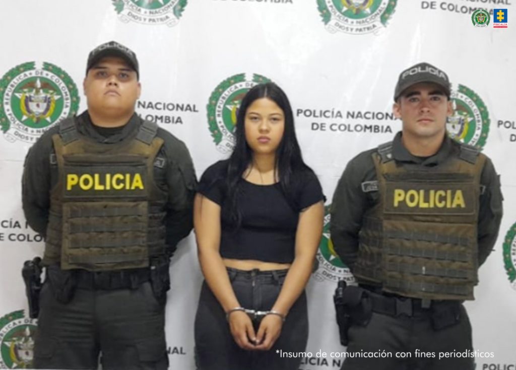 en la imagen se ve una persona detenida bajo custodia de dos integrantes de la Policía Nacional. Detrás, un backing institucional.