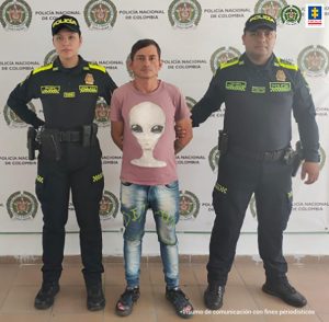 En la imagen se ve una persona detenida bajo custodia de dos uniformados de la Policía Nacional. Detrás suyo un backing institucional.