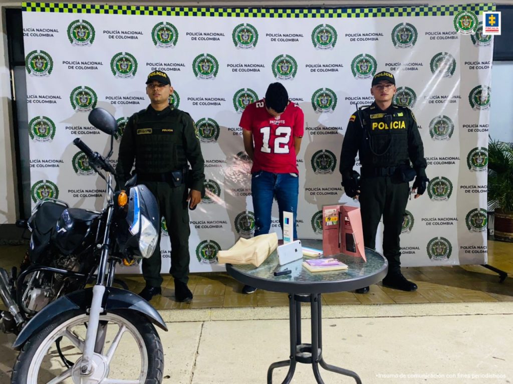 En la imagen se ve una persona detenida en custodia de dos policías. Hay una mesa con elementos recuperados y una motocicleta. Detrás suyo un backing de la institución policial. 