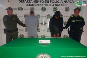  en la imagen se ven dos hombres detenidos. Uno mirando hacia el frente y el otro dando la espalda. Son custodiados por integrantes de la Policía Nacional. Delante suyo una mesa con un celular incautado. 