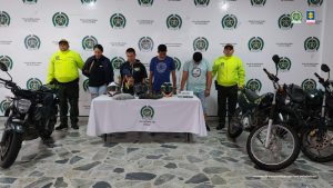 en la imagen se ven cuatro personas detenidas bajo custodia de dos integrantes de la Policía Nacional. Se visualiza igualmente tres motocicletas incautadas y en una mesa varios cascos y documentación. Detrás suyo un backing institucional.