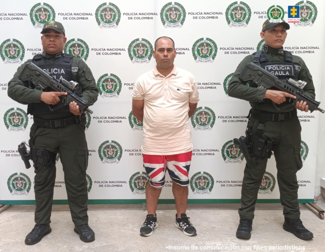 En la imagen se ve una persona detenida bajo custodia de dos uniformados de la Policía Nacional. Detrás suyo un backing institucional.