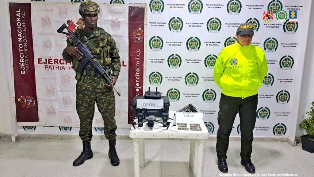 En la imagen se ve una mesa con un dron y otros elementos incautados bajo custodia de un militar y un policial. Detrás suyo backings institucionales.