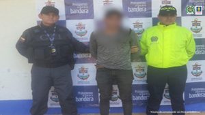 El imputado está en medio de un servidor del CTI de la Fiscalía y un uniformado de la Policía, atrás de ellos hay un pendón institucional.