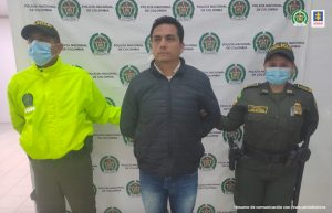 En la imagen se ve una persona detenida en custodia de dos integrantes de la Policía Nacional. Detrás suyo un backing institucional.  