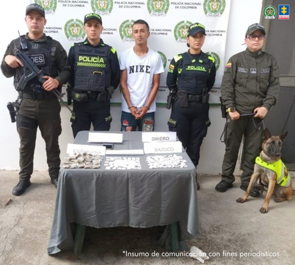 En la imagen se ve una persona detenida bajo custodia de varios uniformados de la Policía Nacional. Frente a ellos una mesa con elementos incautados, y detrás un backing institucional.