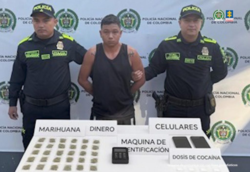 En la imagen se ve una persona detenida bajo custodia de la Policía Nacional. Delante de ellos una mesa con elementos incautados. Detrás, un backing institucional.