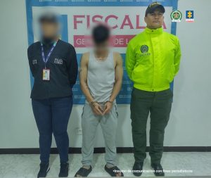 El capturado está de frente custodiado por investigador del CTI de la Fiscalía y Policía. Detrás banner de la Fiscalía.