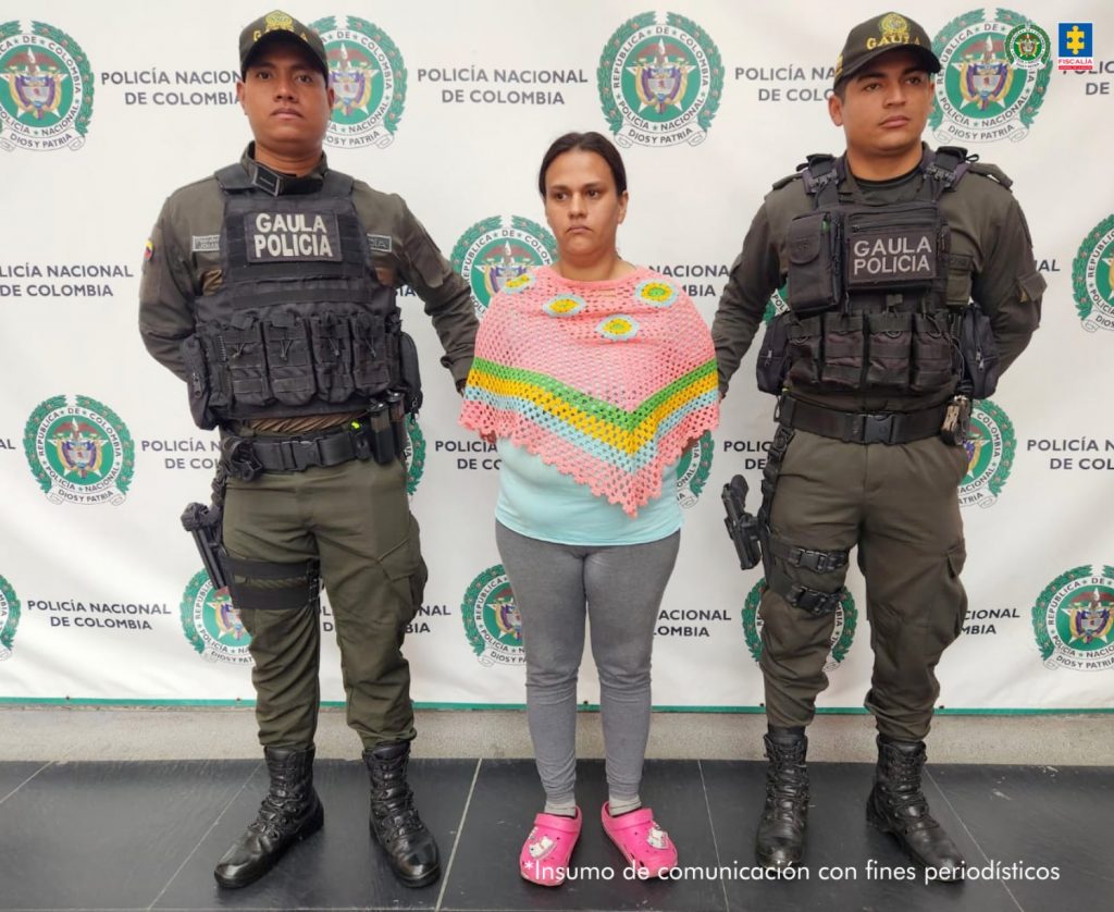 En la imagen se ve una mujer detenida bajo custodia de dos uniformados de la Policía Nacional. Detrás suyo un backing institucional.