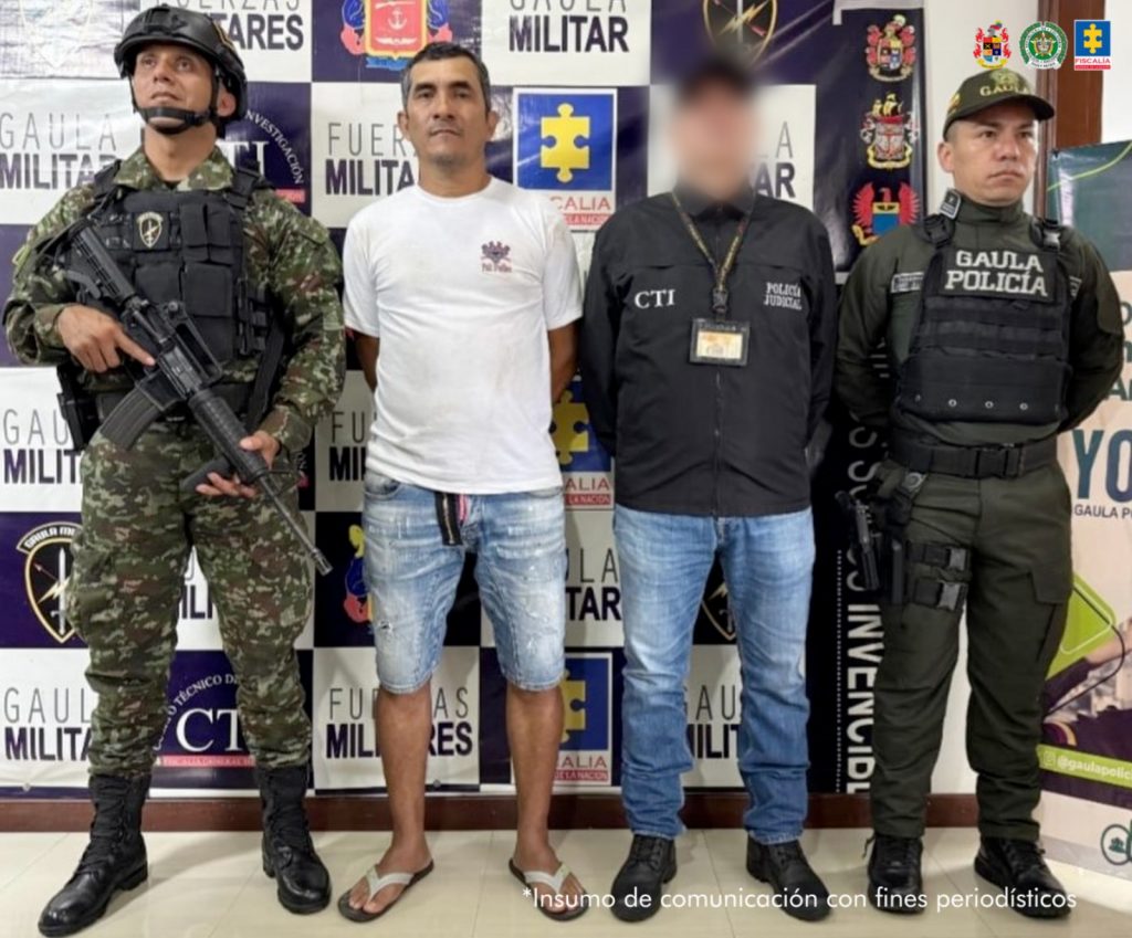 en la imagen se ve una persona detenida bajo custodia de las autoridades. Detrás suyo un backing institucional.