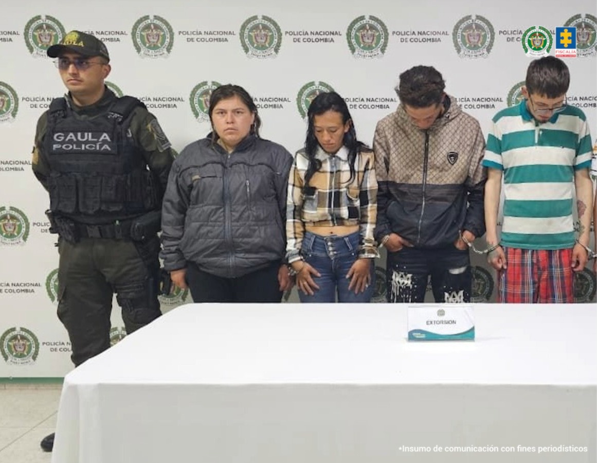 En la imagen se ven cuatro personas detenidas bajo custodia de un uniformado de la Policía Nacional. Detrás suyo un backing institucional.
