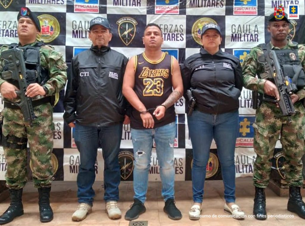 En la fotografía se aprecia a Víctor Alfonso Paz Rendón, de pie y con los brazos adelante y esposado. Viste camisa esqueleto negra con estampado amarillo con el número 23 y la palabra ‘Lakers’. Al lado izquierdo hay una servidora del CTI de la Fiscalía y un uniformado del Ejército Nacional; al lado derecho hay otro uniformado del Ejército. Detrás de ellos hay un pendón institucional con logos de la Fiscalía y del Gaula