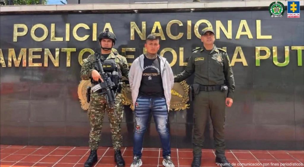 Fotografía de la captura de un hombre en el departamento de Putumayo. En el centro de la imagen aparece el detenido, vestido con una chaqueta gris y un pantalón azul con parches, con las manos hacia atrás. A su lado izquierdo se encuentra un uniformado del Ejército que porta camuflado militar, casco y un fusil. A su lado derecho está un policía uniformado. Los tres posan frente a un muro negro con letras doradas que dicen “Policía Nacional Departamento de Policía Putumayo”. En la parte superior derecha de la imagen están los logos de la Policía Nacional y de la Fiscalía General de la Nación.