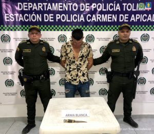 en la imagen se ve una persona detenida bajo custodia de dos uniformados de la Policía. Detrás suyo un backing institucional.