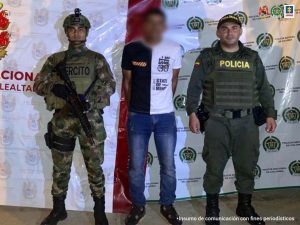 En la imagen se ve una persona detenida bajo custodia de un militar y de un policía. Detrás suyo dos backings de cada una de las instituciones.