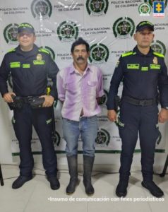 En la imagen se ve una persona detenida bajo custodia de dos uniformados de la Policía Nacional. Detrás, un backing institucional.