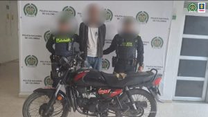 En la fotografía aparece un hombre delgado, alto de pie, con los brazos atrás, vestido con una camiseta blanca, chaqueta gris encima, jean azul, custodiado por dos agentes de la Policía Nacional. Delante de ellos aparece la moto que se le incautó.