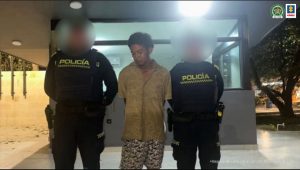 En la fotografía está un hombre de pie, joven, moreno de cabellos negros crespo, con los brazos atrás, vestido de camisa beige con pantaloneta café estampada, acompañado por dos agentes de la Policía Nacional en un CAI de esa institución.