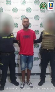  En la imagen esta un hombre de mediana estatura, moreno, de pie, con los brazos atrás, vestido de camiseta roja y bermuda de jean azul. A su lado están dos policías que lo tienen agarrado de sus brazos. Detrás de ellos hay un pendón de esa institución.