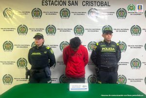 En la imagen se ve una persona detenida bajo custodia de dos integrantes de la Policía Nacional. Detrás suyo un backing institucional.