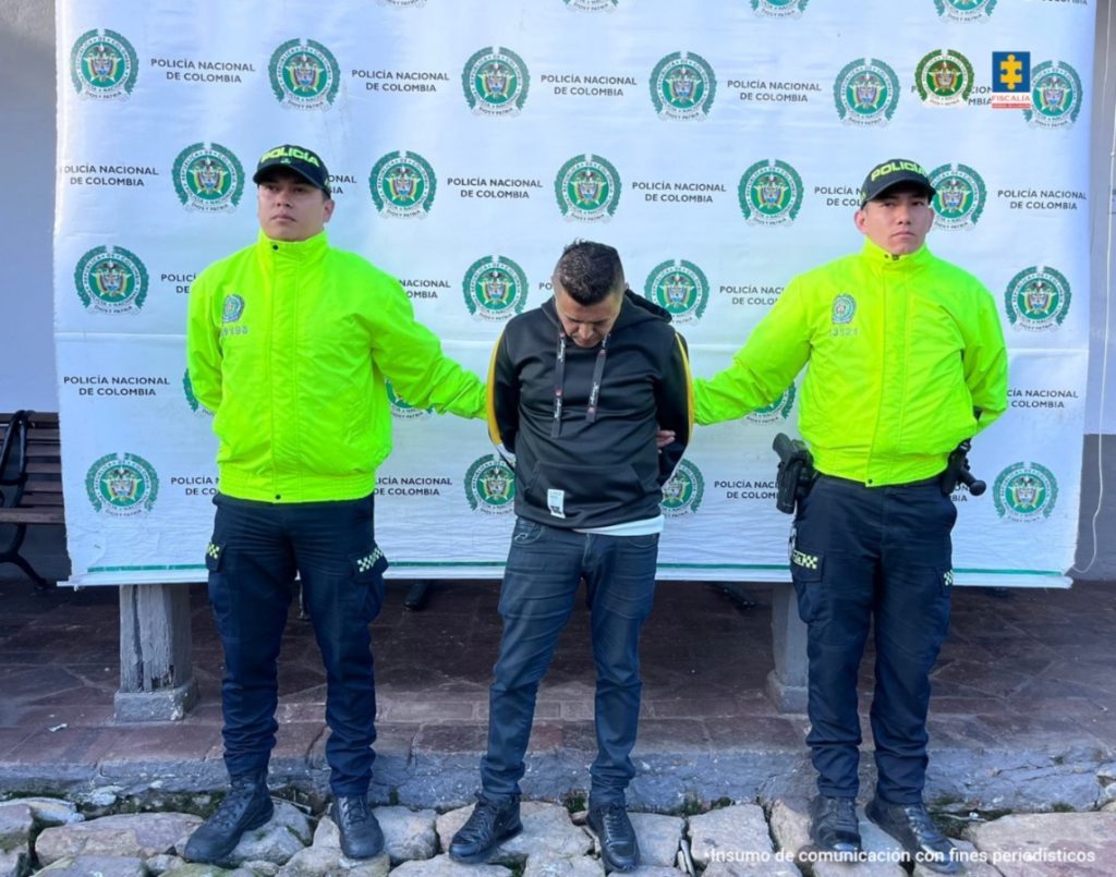 en la imagen se ve una persona detenida bajo custodia de dos integrantes de la Policía Nacional, detrás suyo un backing institucional.