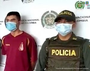 en la imagen se ve una persona detenida bajo custodia de dosuniformados de la Policía Nacional. Detrás suyo un backing institucional.