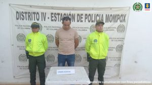 En la imagen se observa a un hombre con la cabeza agachada de camiseta rosada con jean, custodiado por dos agentes de la Policía Nacional delante de un pendón de esta institución.