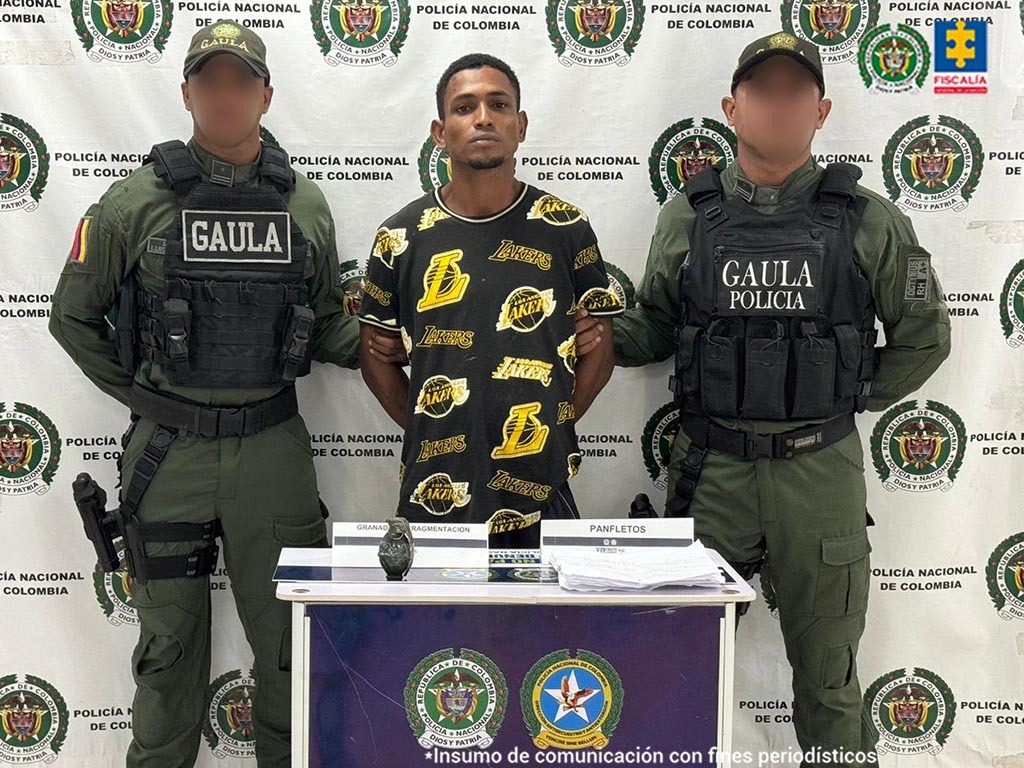 En la fotografía aparece un hombre de mediana estatura, con los brazos atrás, moreno, de cabellos crespos moreno, con barba pequeña, vestido de camiseta negra con amarillo, custodiado por dos uniformados de la Policía Nacional.