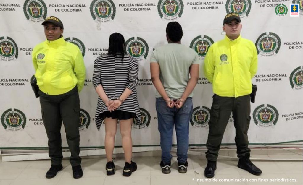 En la imagen se a una pareja de espalda custodiada por dos agentes de la Policía Nacional delante de un pendón de esa institución.