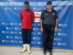 En la imagen aparece un hombre con el rostro difuso vestido de camiseta roja con jean azul y botas blancas, custodiado por un uniformado del CTI. Detrás de ellos hay un pendón azul que dice Fiscalía General de la Nación.