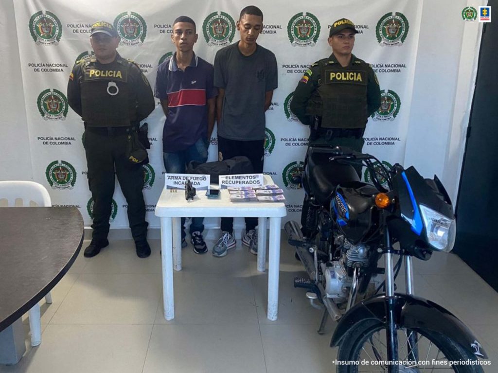 En la fotografía se observa dos hombres con camisetas oscuras custodiado de dos agentes de la Policía Nacional, delante de un pendón de esa institución y detrás de una mesa blanca que en su superficie tiene dinero en efectivo y el arma incautada.