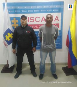 En la fotografía se observa a un hombre moreno de camiseta gris con jean claro, custodiado por un uniformado del CTI y banderas de Colombia y Fiscalía a cada lado. Igualmente se visibiliza detrás de ellos dos un pendón de la Fiscalía General de la Nación.