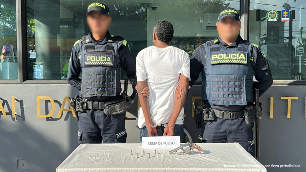 En la fotografía está un hombre de pie, de espalda, con los brazos esposados, cabellos negros crepos, vestido con una camiseta blanca y un jean azul. Se encuentra en las afueras de un CAI custodiado por dos policías. 