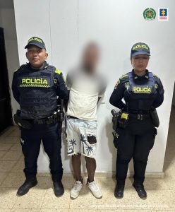 En la imagen hay un hombre con el rostro difuso de pie, vestido con camiseta blanca, bermuda de jean y tenis blanco, custodiado por dos agentes de la Policía Nacional.