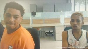 En la imagen están los medios cuerpos de dos hombres morenos, uno de cabellos negro con camiseta naranja y el otro de cabello crespo pintado de amarillo, con camisilla blanca y brazos cruzados. Se encuentran sentados al interior de una oficina asistiendo virtualmente a la audiencia en la que se le procesó.