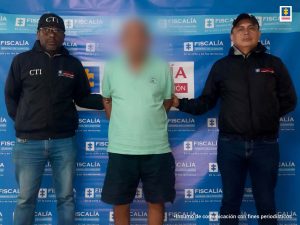 En la imagen está un hombre con el rostro difuso vestido de camiseta verde con bermuda negra, custodiados por dos uniformados del CTI. Detrás de ellos aparece un pendón azul que dice Fiscalía General de la Nación.