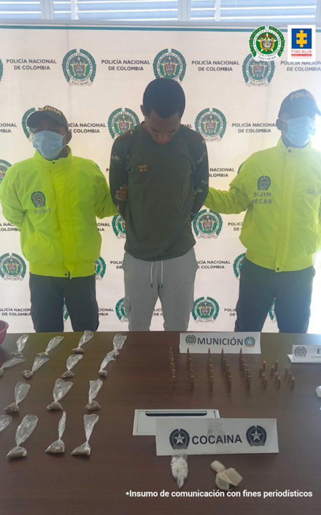 En la imagen se observa a un hombre con los brazos atrás de buzo verde y sudadera gris, custodiado por dos agentes de la Policía Nacional. Delante de él se ve una mesa con la droga y las municiones incautadas.
