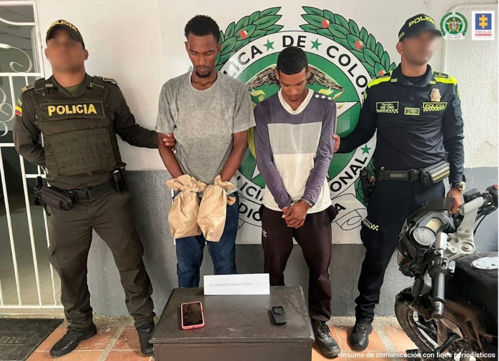 En la fotografía aparecen dos hombres vestidos con camisetas grises, uno con jean y el otro con sudadera, custodiados por dos agentes de la Policía Nacional. Ellos están ubicados delante de una pared que tiene el logo de esa institución. También se encuentra una mesa con dos celulares y una motocicleta.