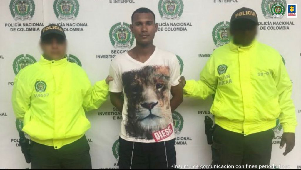 En la imagen aparece un hombre de tez morena, vestido con una camiseta blanca que tiene estampada la cara de un tigre y bermuda blanca, custodiado por dos uniformados de la Policía Nacional. Detrás de ellos hay un pendón de la Policía Nacional.