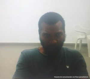 En la fotografía aparece el rostro de un hombre moreno, ojos grandes, nariz gruesa, cabellos negros crespo y barba. Se encuentra vestido con una camiseta negra y está sentado escuchado la audiencia en la que se le procesa.