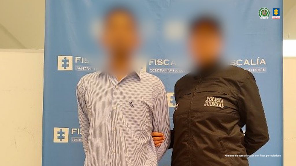 En la fotografía aparece de pie el presunto implicado, con los brazos atrás. Viste camisa blanca con estampado, pantalón gris oscuro y zapatos de color café claro. Al extremo izquierdo de él hay una servidora del CTI de la Fiscalía. Detrás de ellos hay un pendón institucional de la Fiscalía.