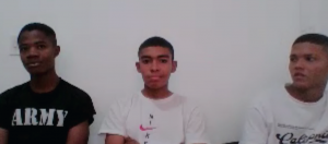 n la imagen aparecen tres jóvenes sentados uno al lado del otro frente a una pared blanca, como si estuvieran en un espacio interior sencillo.El joven de la izquierda tiene el cabello corto y viste una camiseta negra con la palabra “ARMY” en letras grandes. Mira hacia el frente con una expresión neutral.  El joven del centro tiene el cabello corto y lleva una camiseta blanca con un pequeño diseño en el pecho. Está mirando directamente a la cámara y parece tener los brazos cruzados.  El joven de la derecha también viste una camiseta blanca con un texto o diseño en el frente. Su cabeza está ligeramente girada hacia el centro, como si estuviera mirando al joven que está a su lado.