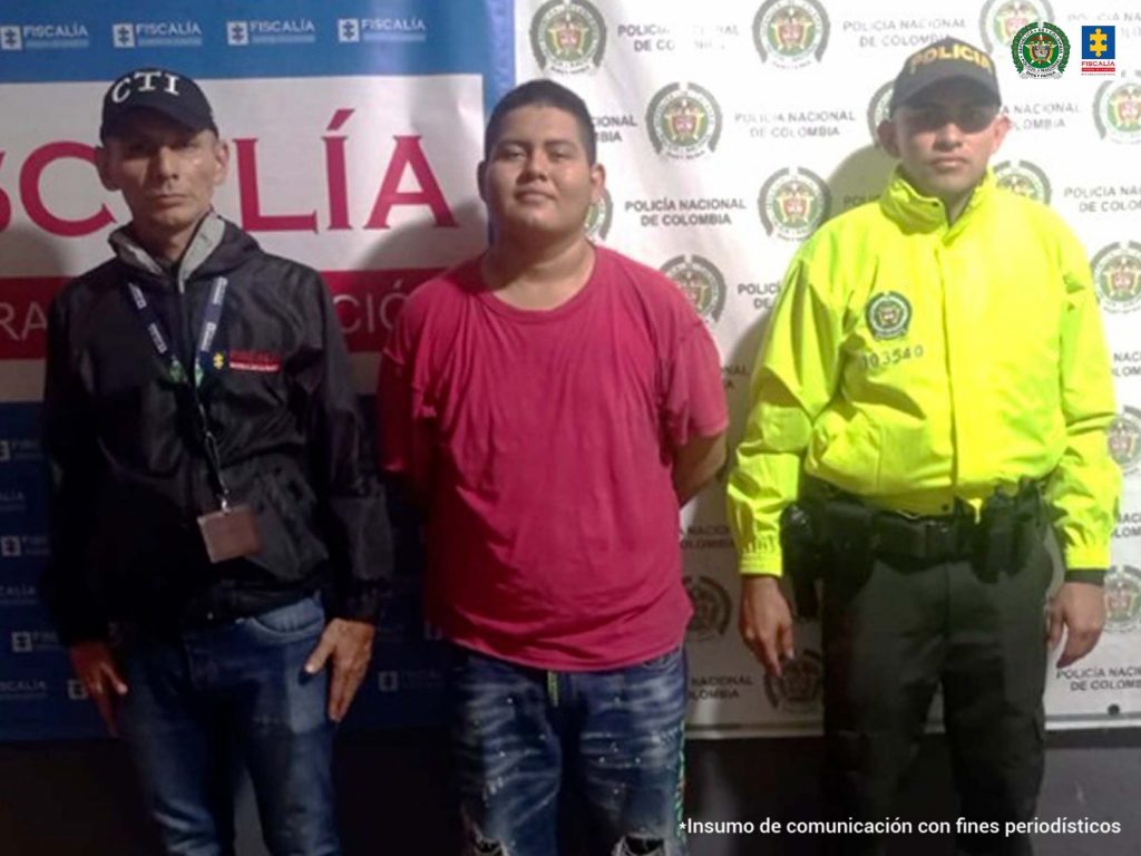 en la imagen se ve una persona detenida en custodia de integrantes del CTI de la Fiscalía y de la Policía Nacional. Detrás suyo un backing de ambas instituciones.