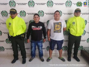  En la imagen se ven dos personas detenidas bajo custodia de dos integrantes de la Policía Nacional. Detrás suyo un backing de la institución.