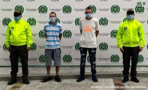 en la imagen se ven dos personas detenidas bajo custodia de dos integrantes de la Policía Nacional. Detrás suyo un backing de la misma institución.