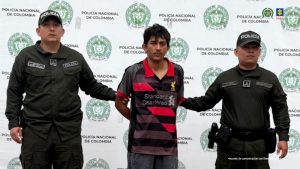 En la imagen se ve un hombre detenido bajo custodia de dos uniformados de la Policía Nacional. Detrás suyo, un backing institucional.
