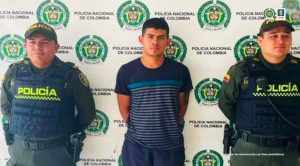 En la imagen se ve una persona detenida bajo custodia de dos integrantes de la Policía Nacional. Detrás suyo un backing institucional.  