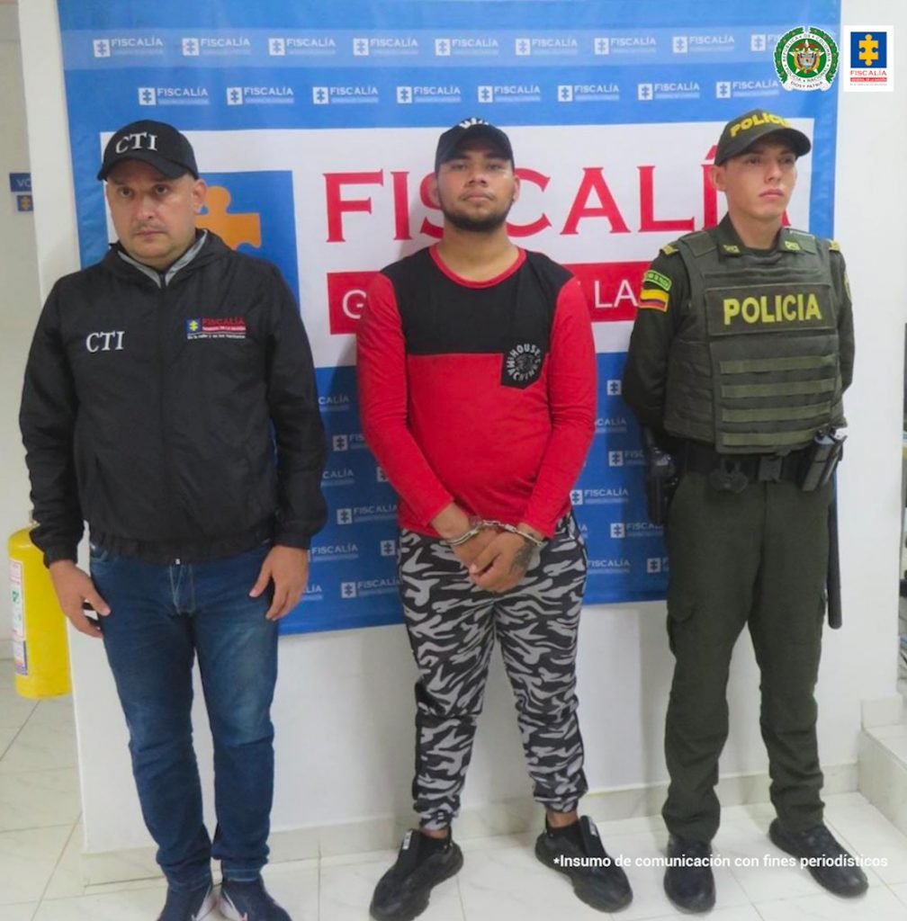 En la imagen se ve una persona detenida bajo custodia de un integrante de la Policía Nacional y del CTI. Detrás suyo un backing de la Fiscalía.